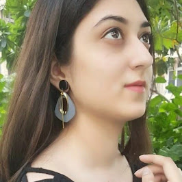A young woman with long dark hair and elegant earrings gazes thoughtfully upwards, surrounded by greenery.